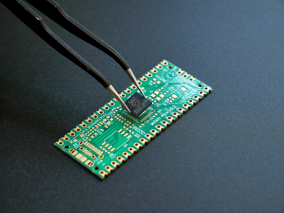 Tweezers hold an RP2040 microcontroller above a green printed circuit board.